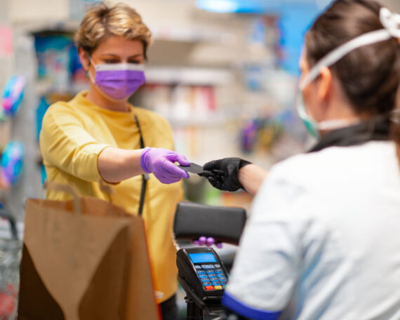 Cashier returning credit card at the cash register to woman with wallet wearing protective face mask and gloves to prevent viruses