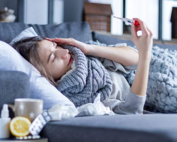 Sick exhausted girl in scarf is lying in bed wrapped in blanket. Young woman with fever and headache is measuring temperature with thermometer, treated at home. Winter cold and flu concept.